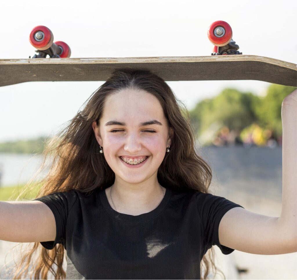 Teenage girl with brown hair smiling with braces while holding a skateboard on her head
