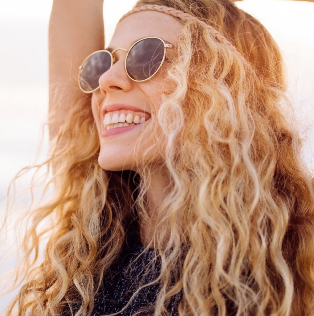 Young female dressed with curly blonde hair smiling on the beach outside in summer